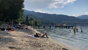 Uma praia à beira do lago com pessoas tomando sol e nadando, um píer se estendendo na água e montanhas ao fundo.