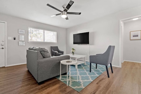 A modern living room with a grey sofa, two armchairs, a small white table, a ceiling fan, a TV, and a window with blinds.