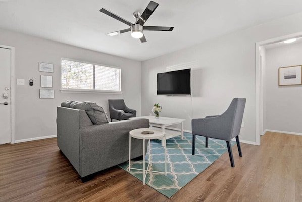 Un salon moderne avec un canapé gris, deux fauteuils, une petite table blanche, un ventilateur de plafond, un téléviseur et une fenêtre avec des stores.