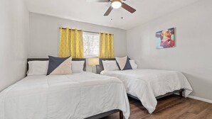 Habitación de hotel con dos camas, un ventilador de techo, una ventana con cortinas amarillas y un cuadro en la pared.