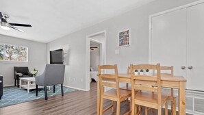 Una sala de estar moderna con una mesa de comedor, sillas, un sillón gris, una mesita blanca y un ventilador de techo.