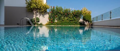 A clear swimming pool with a metal ladder, surrounded by greenery and a clear blue sky.
