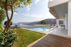 A modern outdoor pool area with a wooden deck, a seating area with chairs and tables, and a view of the sea and hills in the background.