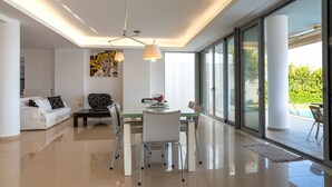 A modern living room with a glass dining table, white sofas, and a large window offering a view of the outdoors. - Blue Oasis Luxury Villa (Malevizi)