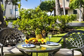 Mesa de comedor exterior con superficie de vidrio, rodeada de sillas, dispuesta con platos, cubiertos, una botella y frutas tropicales.