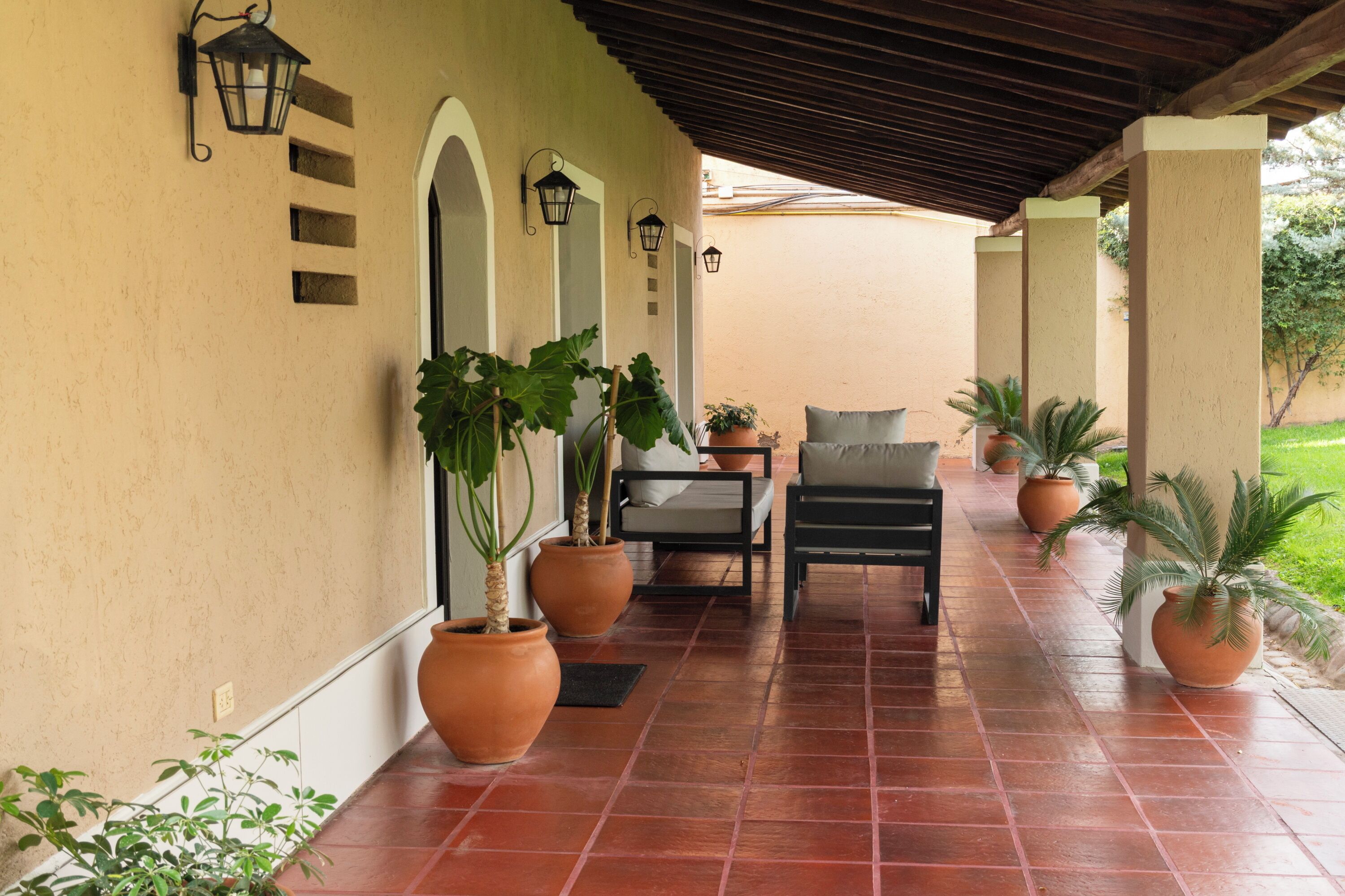 A covered patio area with terracotta tiles, potted plants, a seating area with a chair and table, and wall-mounted lanterns.
