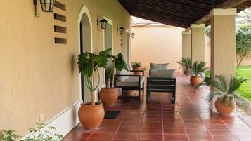 A covered patio area with terracotta tiles, potted plants, a seating area with a chair and table, and wall-mounted lanterns.