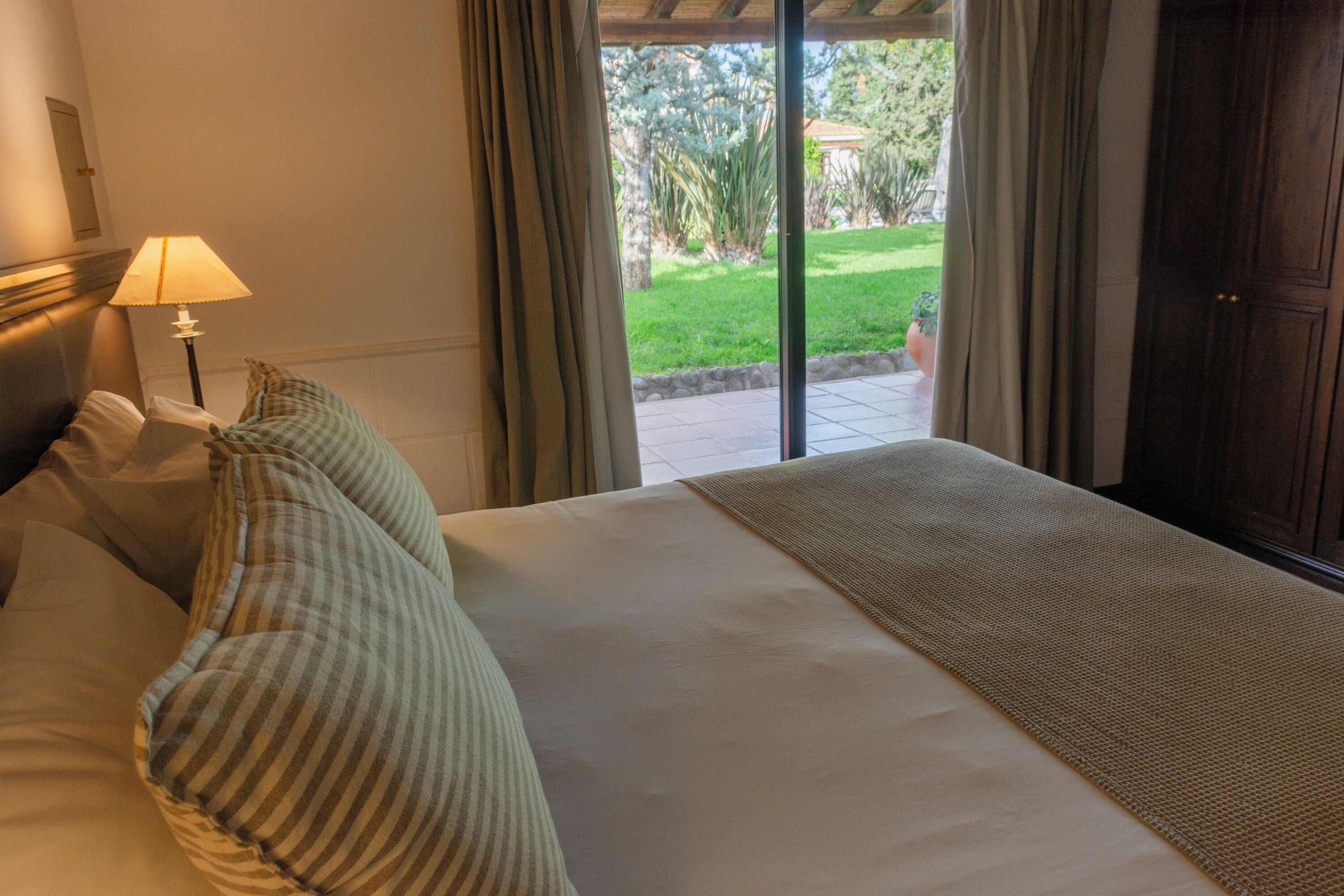 A hotel room with a bed, pillows, a lamp, and a view of a garden through a glass door.