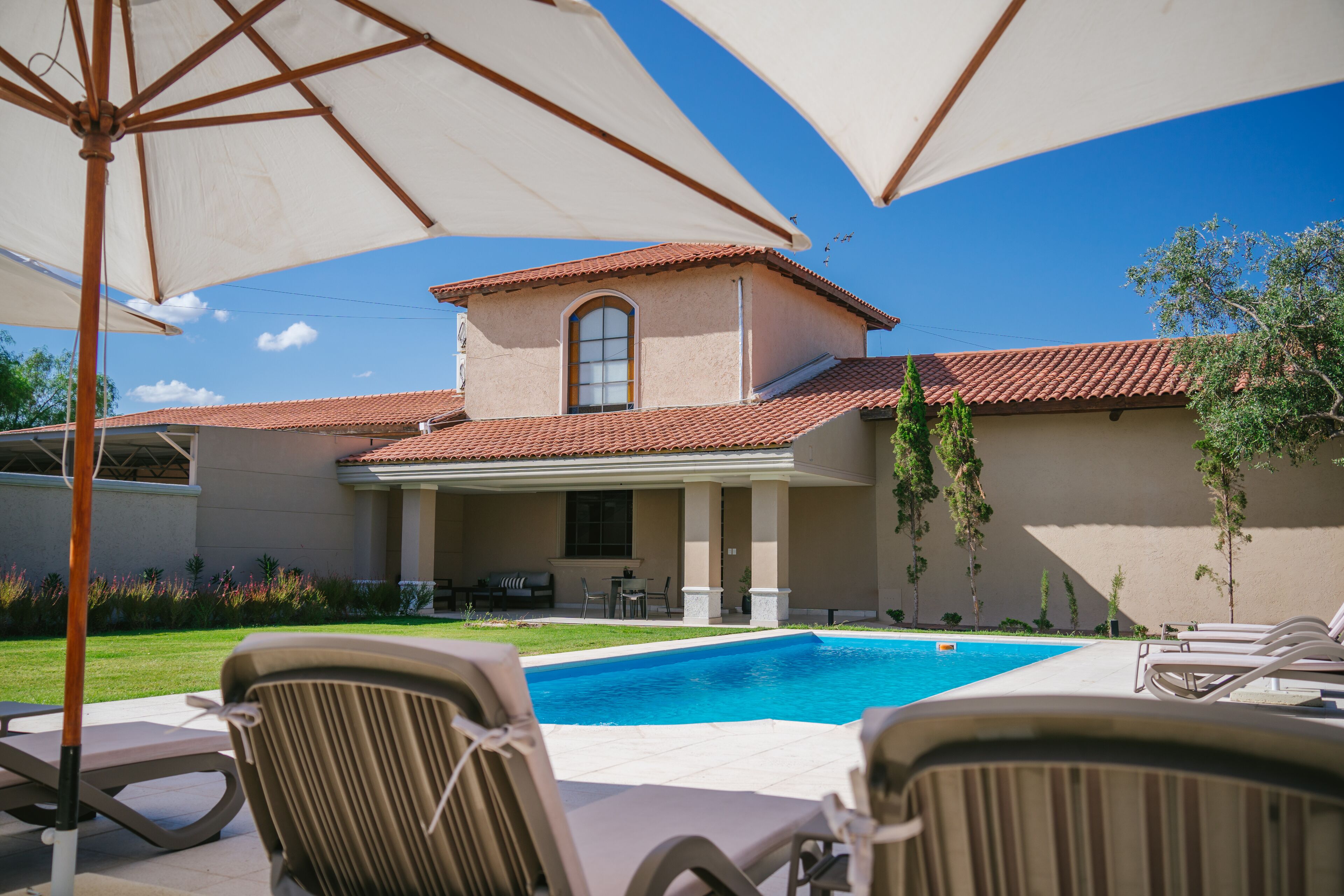 A pool area with lounge chairs and umbrellas, a covered patio, and a building with a tiled roof.