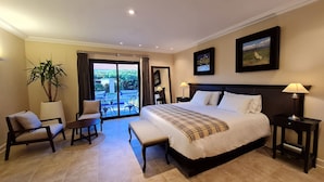 A hotel bedroom with a large bed, two nightstands, a chair, a table, a potted plant, and a balcony with a view of greenery.