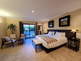 Un dormitorio de hotel con una cama grande, dos mesitas de noche, una silla, una mesa, una planta en maceta y un balcón con vistas a zonas verdes.