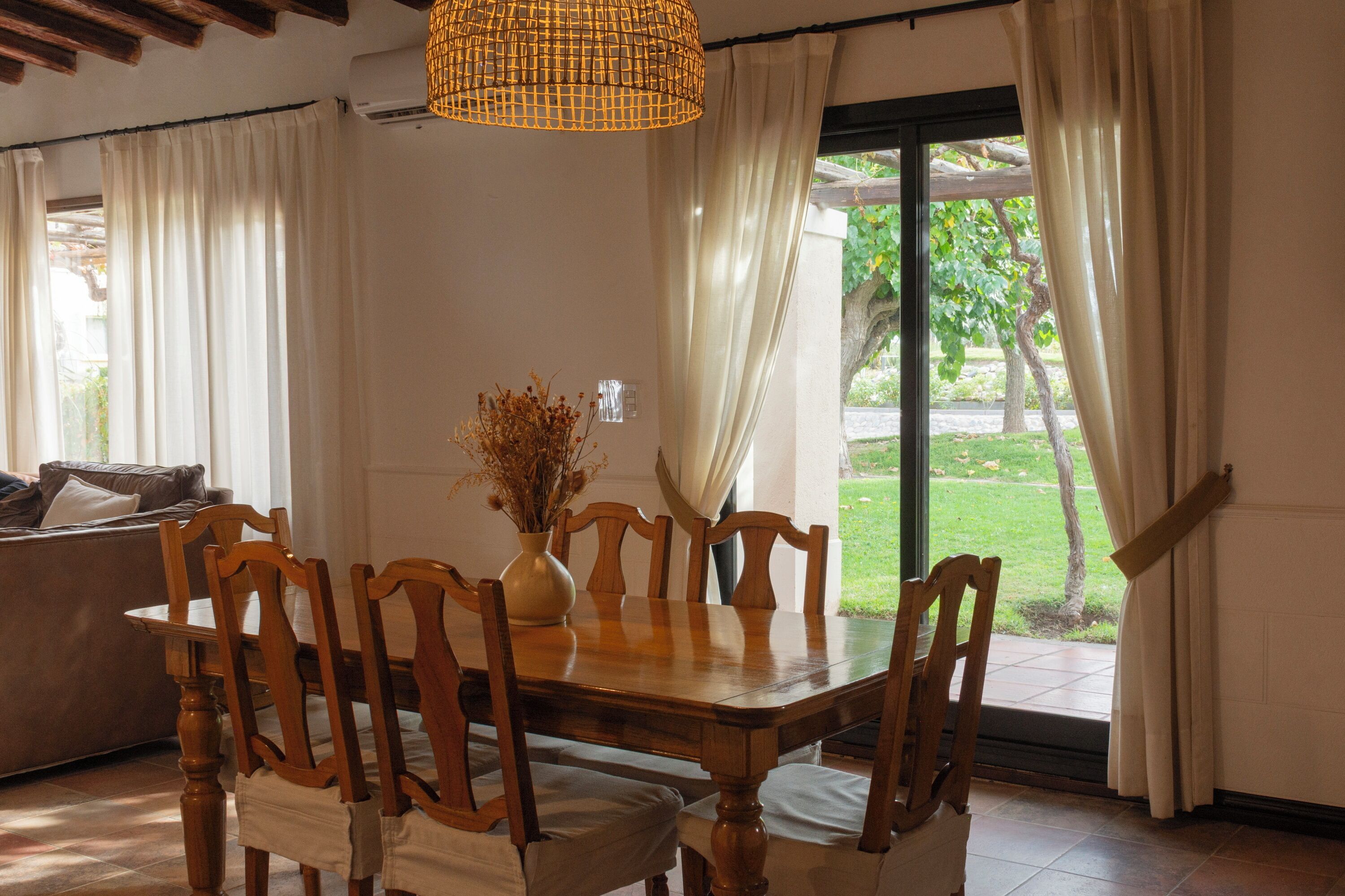 A dining area with a wooden table and chairs, a vase with dried flowers, and a view of a garden through a glass door.