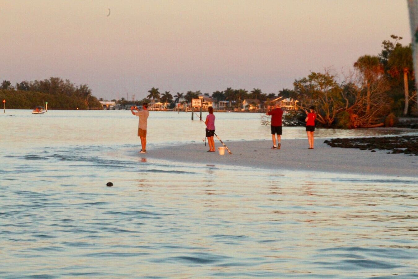 En strandscene med mennesker i vandkanten, en båd i det fjerne og bygninger i baggrunden.