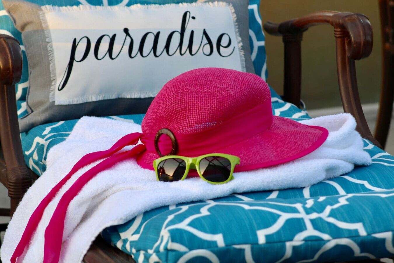 A chair with a blue and white patterned cushion, a pink hat, and a pair of green sunglasses on a white towel.