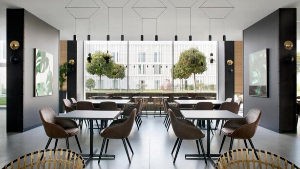 Un comedor moderno con mesas y sillas, diseño geométrico en el techo y amplias ventanas con vistas a árboles.