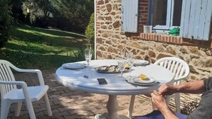 Ein Steinhaus mit Fenster und Fensterläden, ein runder Tisch mit Stühlen und einem Teller Essen.