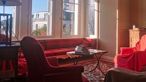 Ein Zimmer mit einem roten Sofa, einem kleinen Tisch mit Büchern, einer Lampe und großen Fenstern mit Blick auf Gebäude.