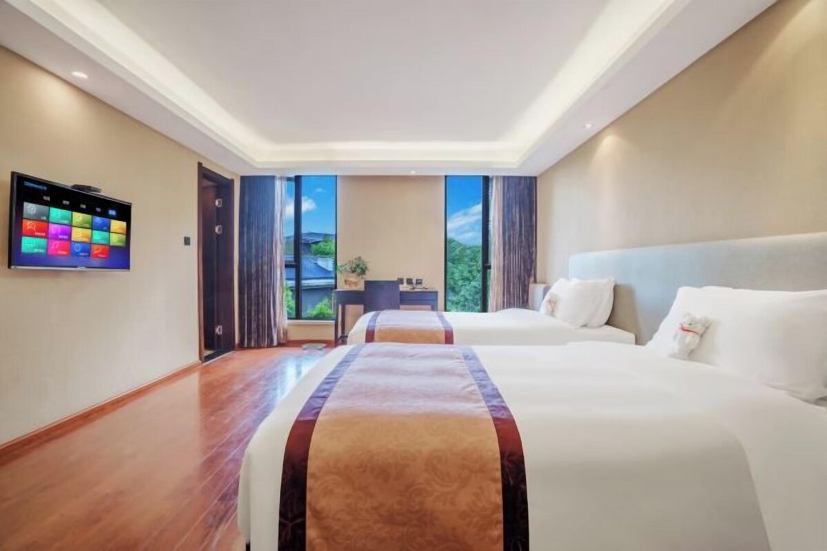 Habitació d'hotel amb dos llits, un televisor de pantalla plana i vista a vegetació a través de grans finestres.