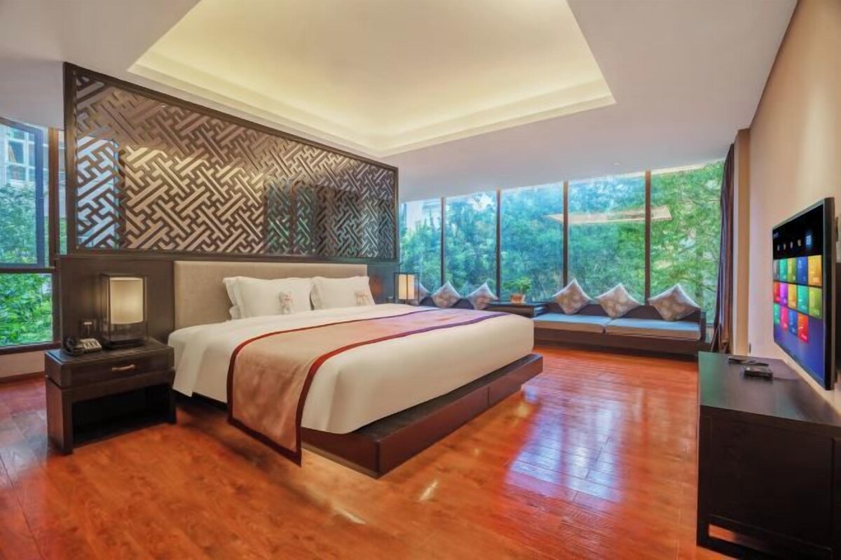 Camera moderna con un letto grande, un comodino, una TV e vista sul verde attraverso ampie finestre.