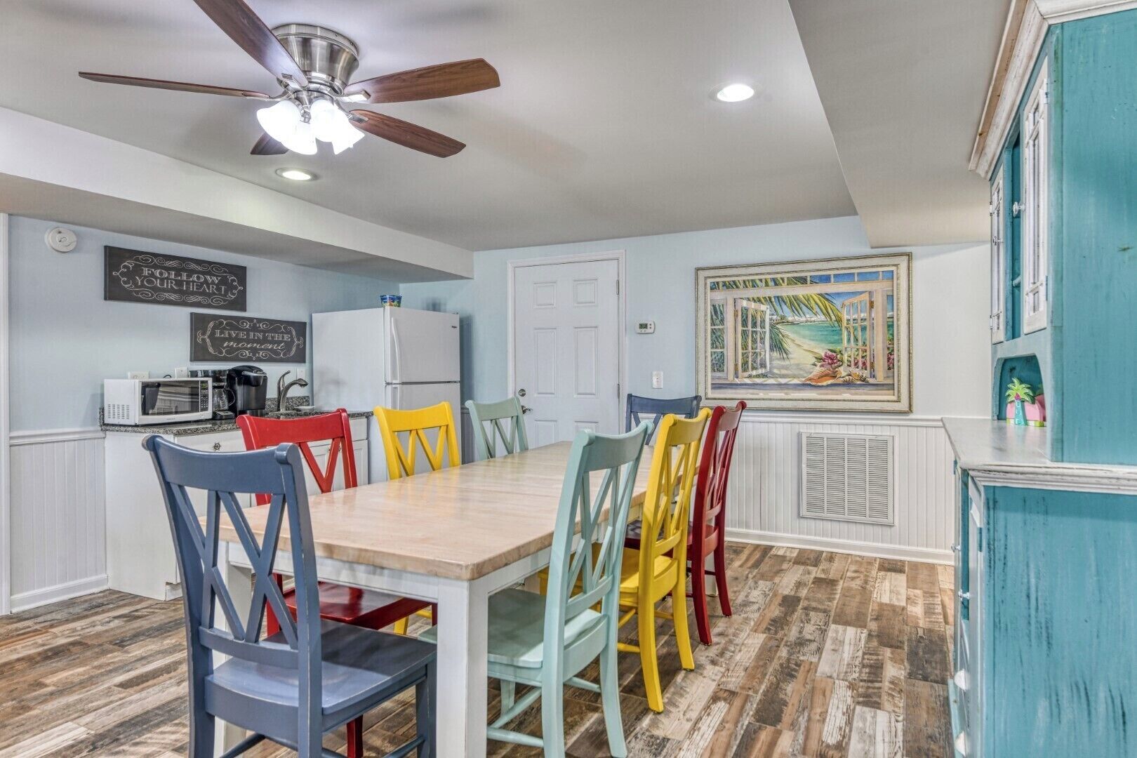 A dining area with a wooden table and colorful chairs, a ceiling fan, a white refrigerator, and a painting on the wall.
