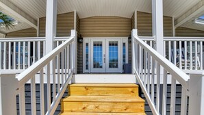 Eine Holztreppe führt zu einer Laube mit weißen Geländern und Blick auf ein Haus mit blauer Tür und Fenstern.