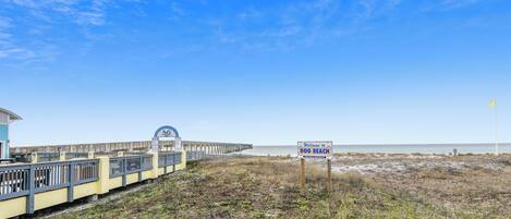 A beach area with a wooden boardwalk, a sign reading "Welcome to Dog Beach," and a pier in the background.