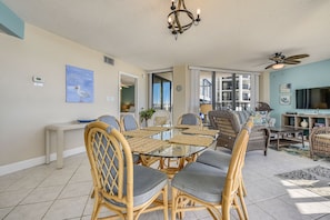 A spacious living area with a glass-top dining table surrounded by wicker chairs, a ceiling fan, and a large window offering a view of the outside.