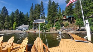 A wooden deck with Adirondack chairs, a dock extending into a lake, an American flag, and a cabin in the background. - Beautiful Log Cabin w/ Beachfront & Stunning Lake Views in Coeur d'Alene Idaho. (Harrison)