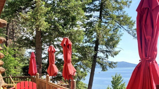 A wooden deck with red Adirondack chairs and umbrellas, overlooking a lake with trees and mountains in the background. - Beautiful Log Cabin w/ Beachfront & Stunning Lake Views in Coeur d'Alene Idaho. (Harrison)