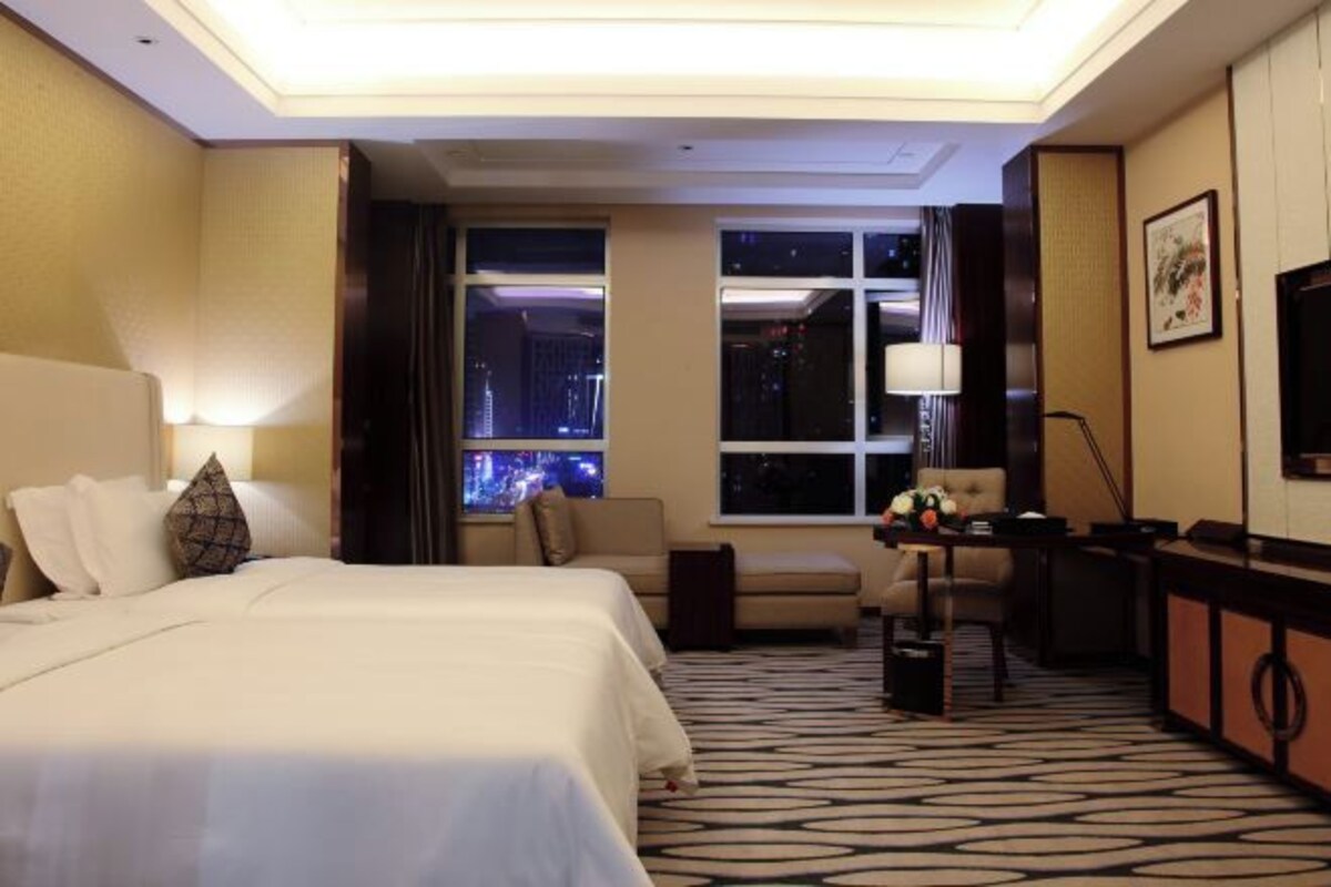 Habitación de hotel con una cama grande, un sofá, un escritorio y un televisor. Se ve una ventana con vistas a la ciudad de noche.