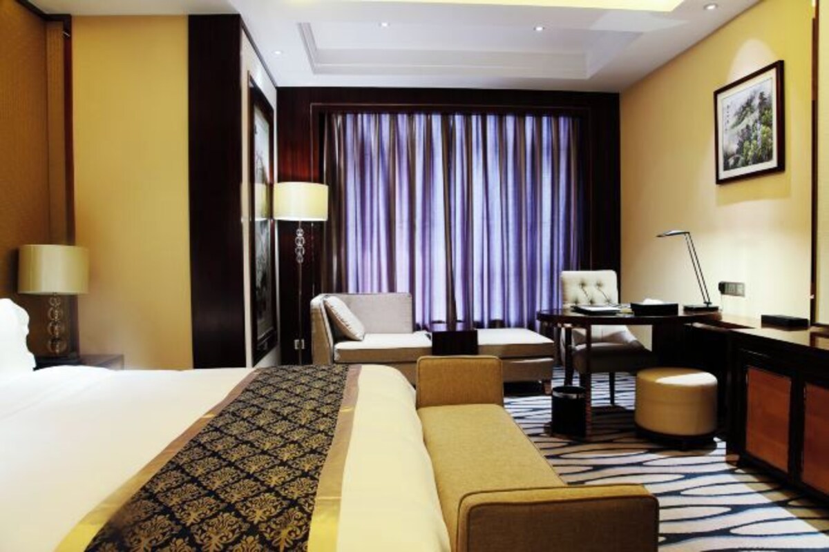 Habitación de hotel con una cama grande, un sofá, un escritorio y una silla. Cuenta con una ventana con cortinas, una lámpara y un cuadro en la pared.