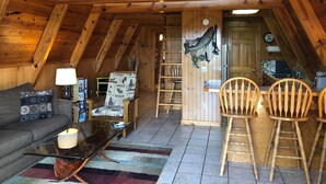 A cozy attic room with wooden beams, a sofa, a glass coffee table, and a dining area with chairs. - Rivers Bend Cabin, Stunning Caddo River & Ouachita Mountain Views (Amity)