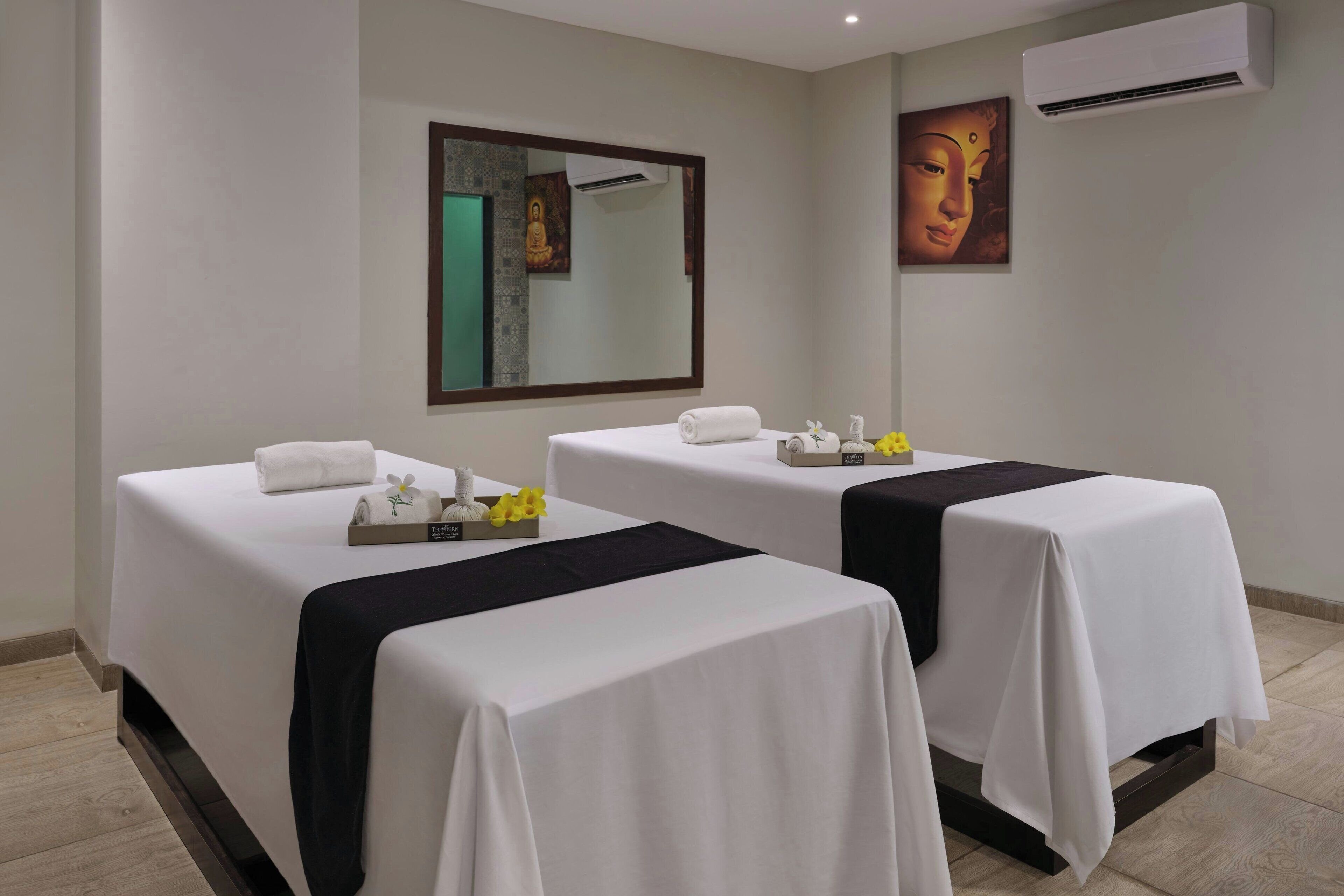 A hotel room with two beds, each covered with white linens and a black runner. There is a mirror above the beds, and a Buddha statue is visible on the wall.