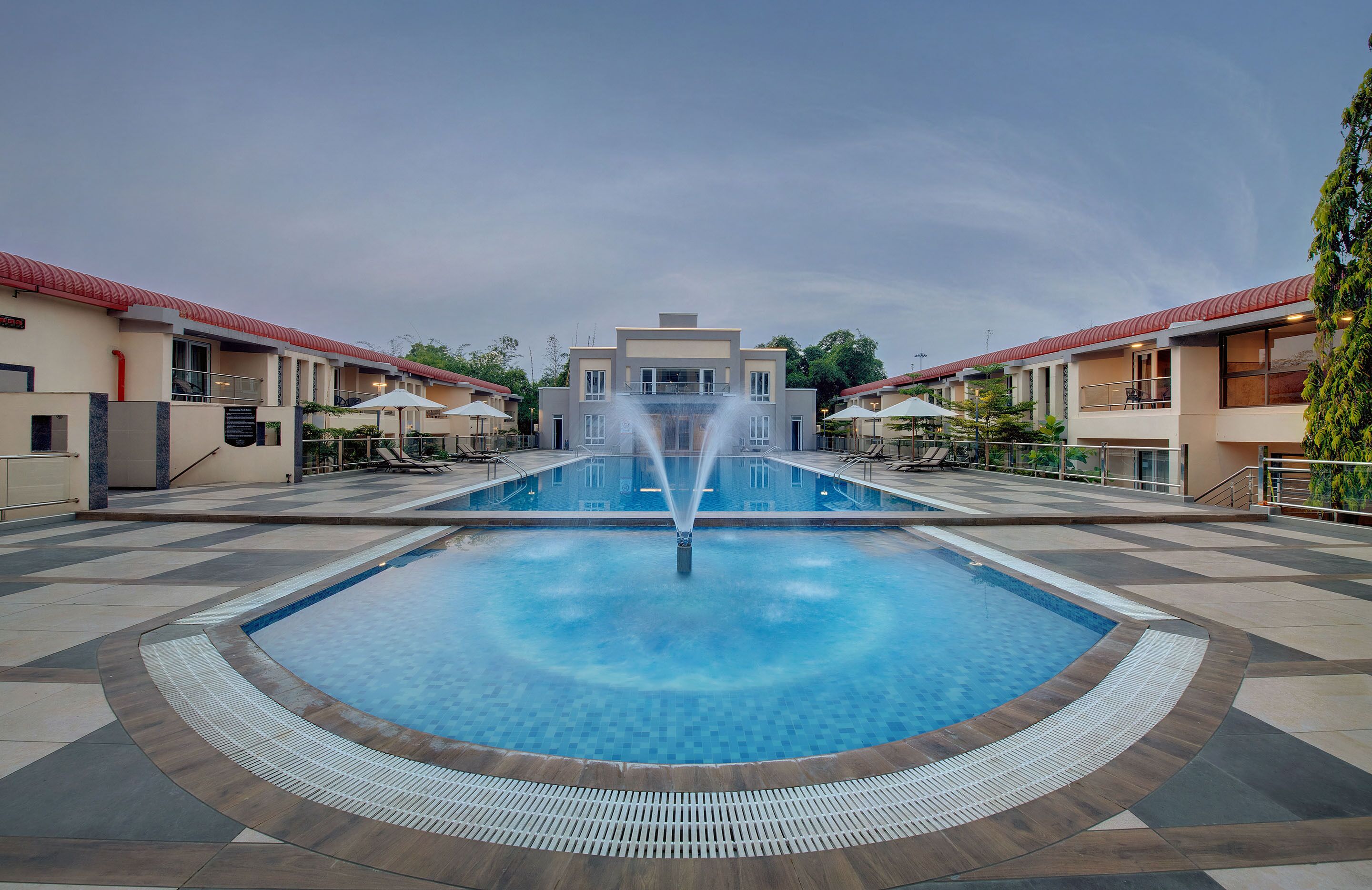 Una piscina con una fuente central, rodeada por una solana empavedada y edificios cercanos con balcones.