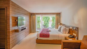 A hotel room with a wooden accent wall, a flat-screen TV mounted above a wooden shelf, a bed with white bedding and a red throw, a bedside table with a lamp, and a window with curtains. - The Fern Sardar Sarovar Resort, Series by Marriott (Garudeshwar)