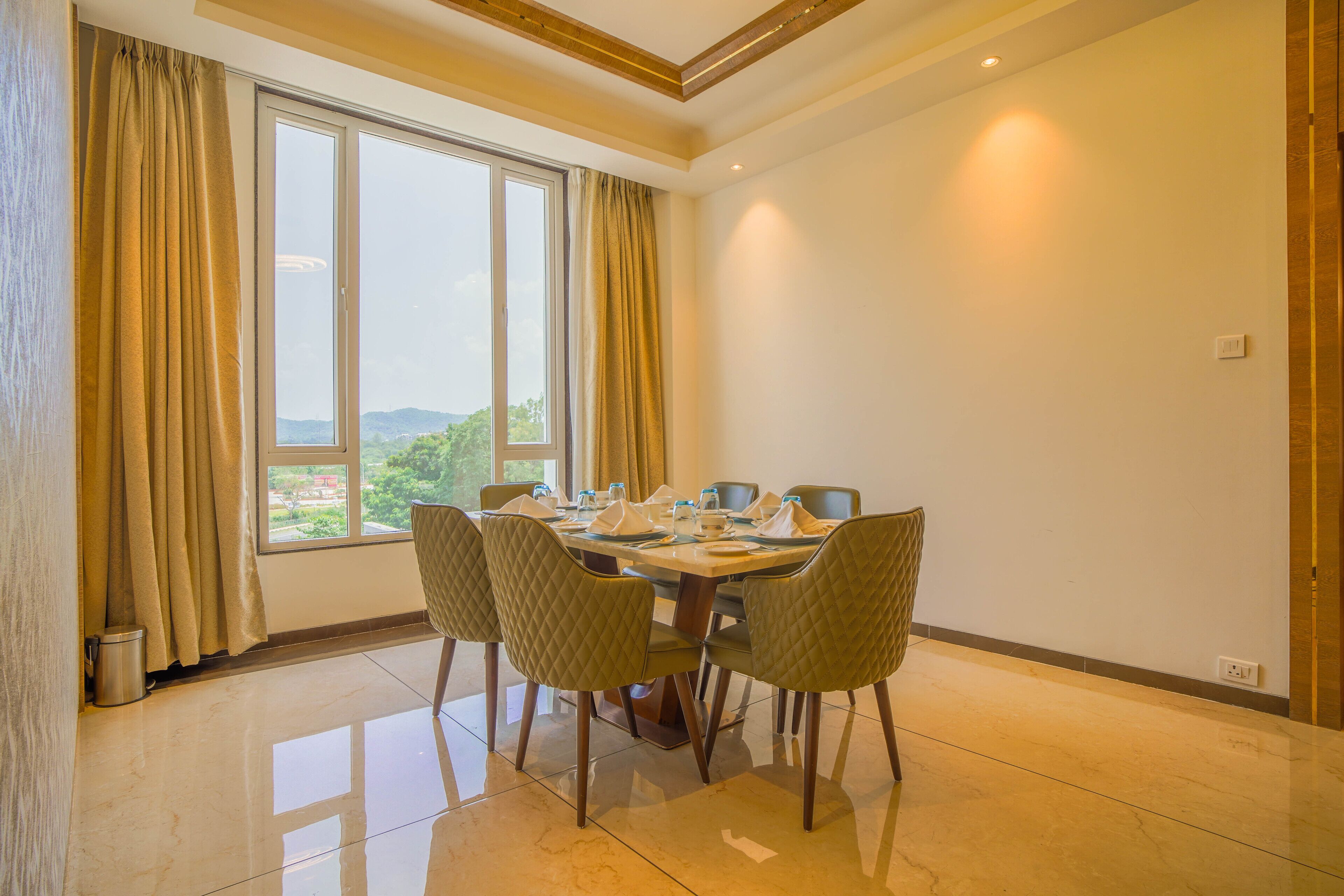 A dining area with a table set for four, a window with curtains, and a view of greenery outside.