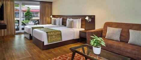 A hotel room with two beds, a sofa, a coffee table, and a balcony with a view of a building and a tree.