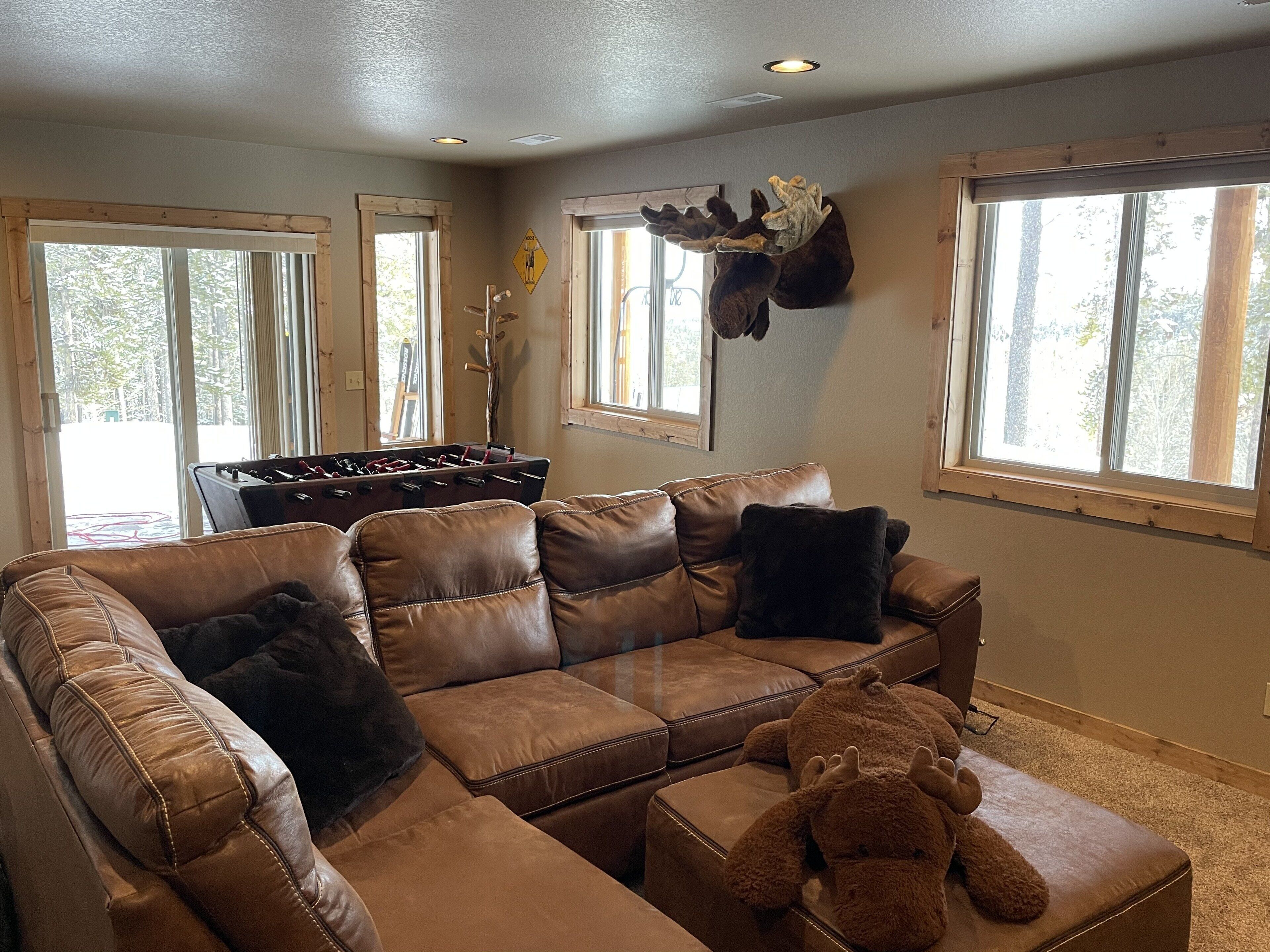 A cozy living room with a brown leather sectional sofa, a foosball table, and a stuffed animal on an ottoman.