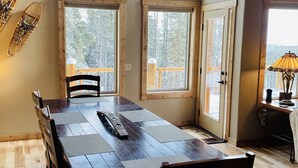 A dining area with a long wooden table, multiple chairs, and large windows offering a view of the outdoors.