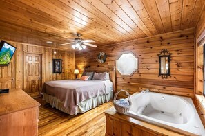 A wooden cabin interior with a bed, a large window, a ceiling fan, and a bathtub.