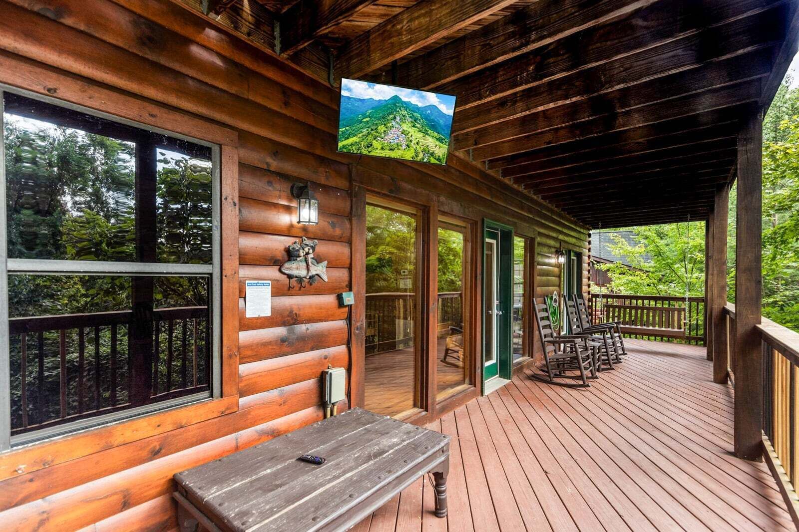Een houten chalet met een overdekte veranda, een bankje en een televisiescherm waarop een landschap te zien is.