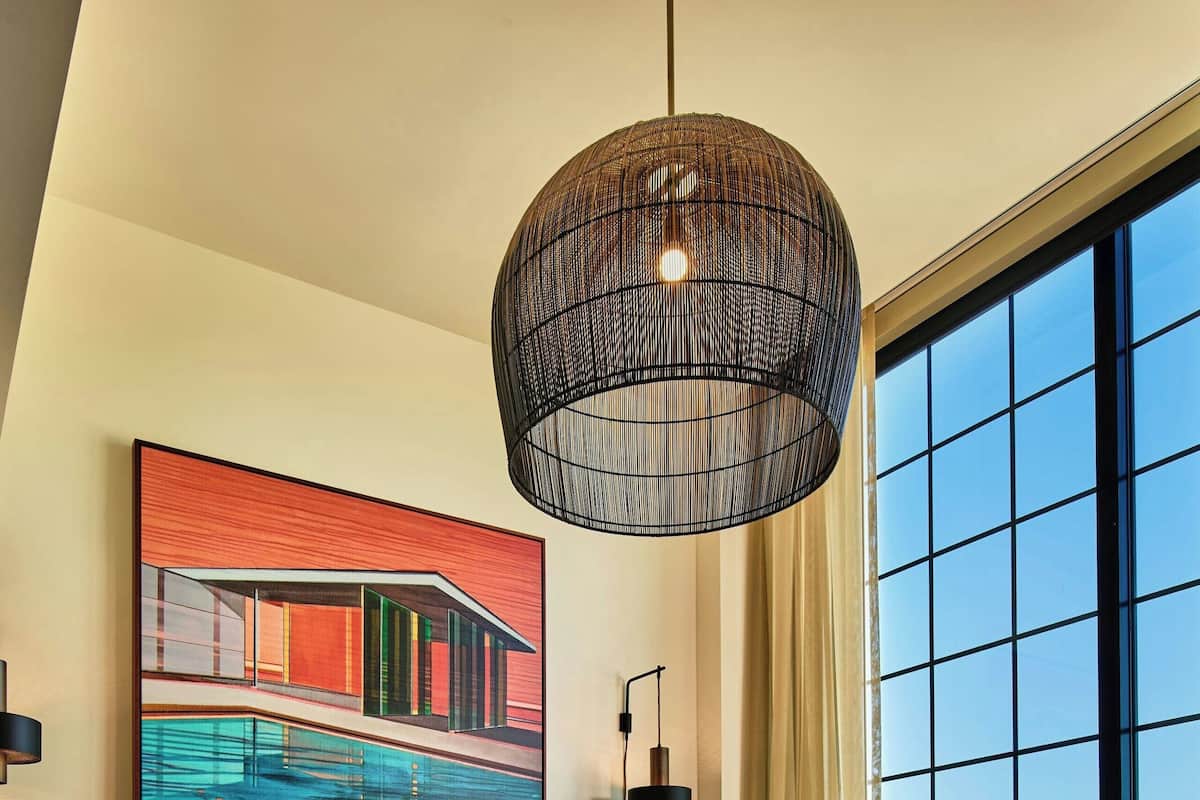 A hotel room with a large bed, a hanging light fixture, a painting, and a view of the city through large windows.
