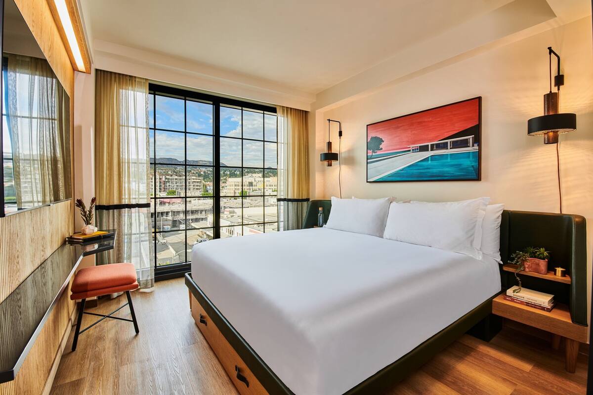 A hotel room with a large bed, a desk with a chair, a small stool, a nightstand with a lamp, and a painting on the wall.