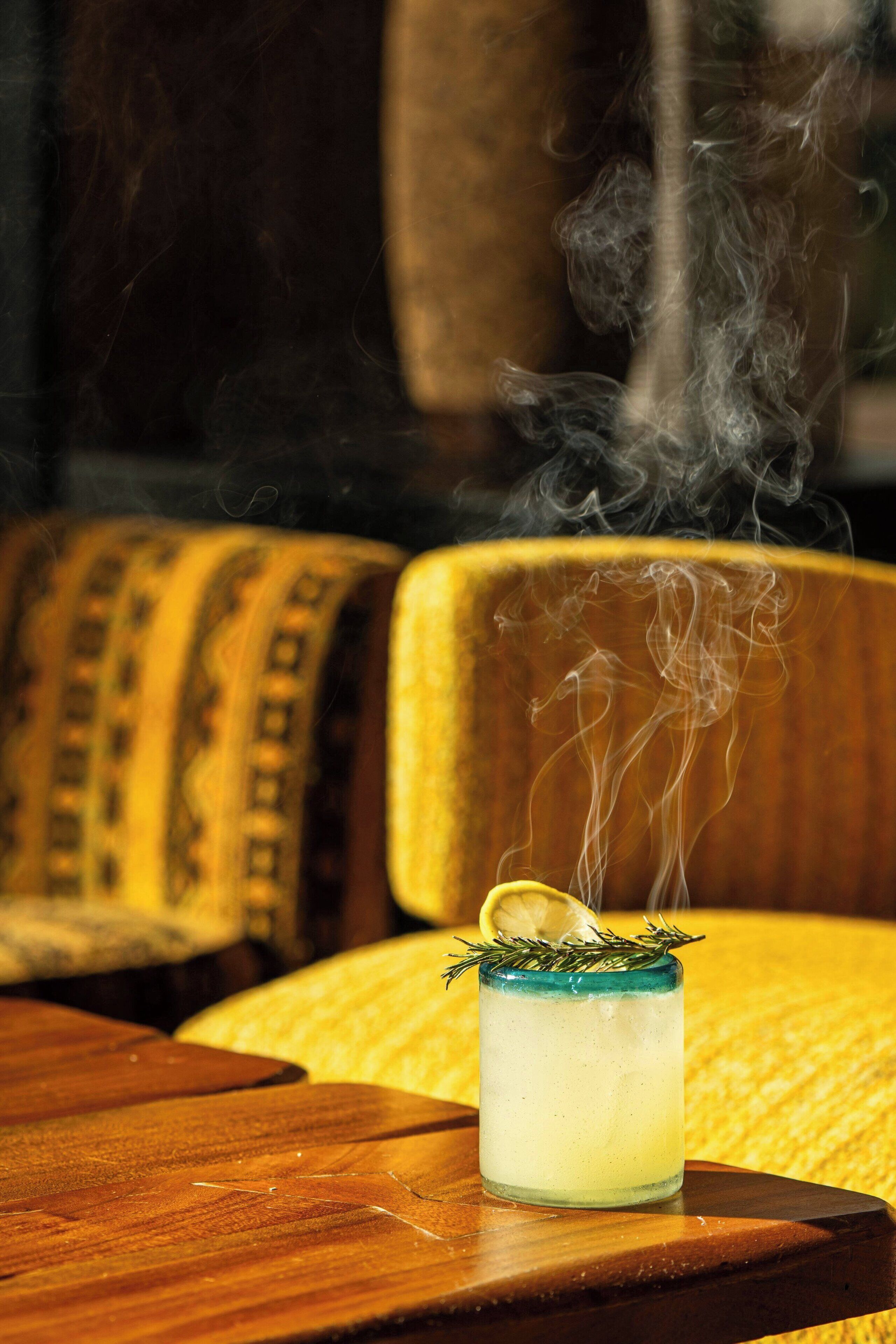 A cocktail with a lemon slice and a sprig of rosemary on a wooden table.