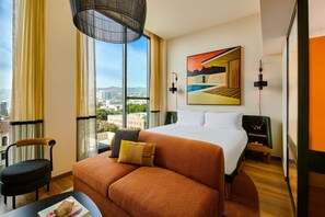 A hotel room with a large bed, a sofa, a chair, a desk, and a view of the city through large windows.