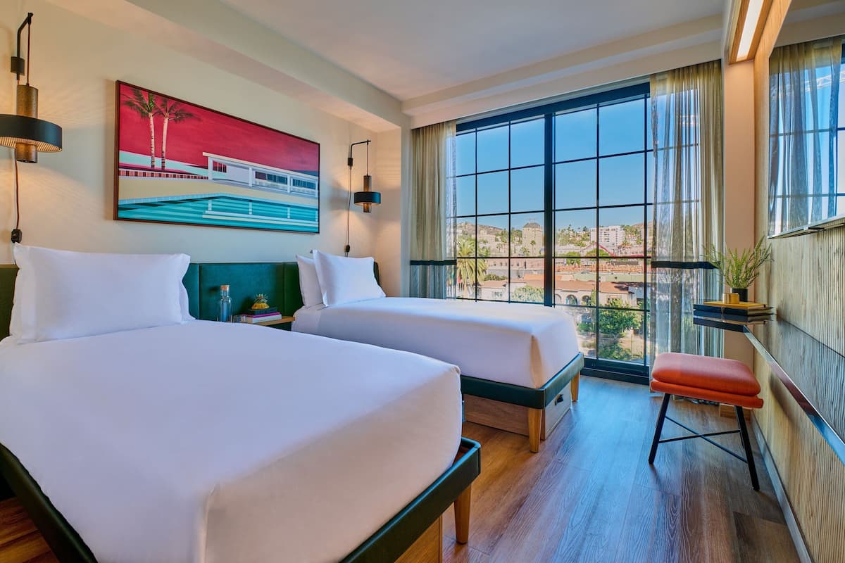 A hotel room with two beds, a large window with a view, a painting on the wall, and a small stool.
