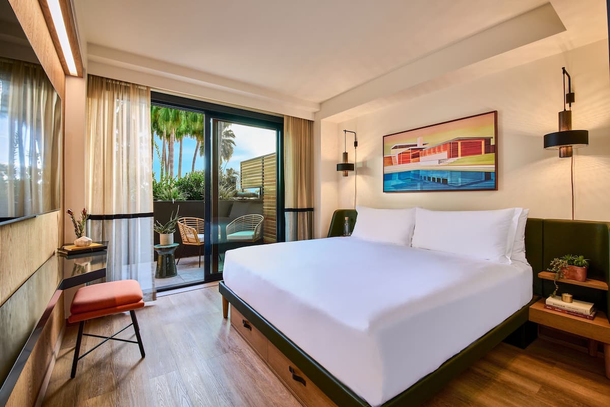 A hotel room with a large bed, a desk with a chair, a small table with a plant, and a sliding glass door leading to a balcony with a view of palm trees.