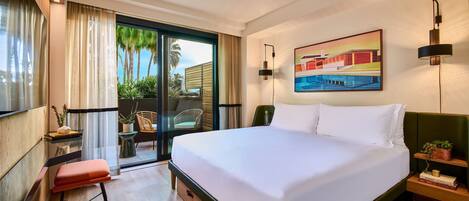 A hotel room with a large bed, a desk with a chair, a small table with a plant, and a sliding glass door leading to a balcony with a view of palm trees.
