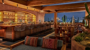 A bar area with wooden beams, a bar counter, and seating. There are plants and a city view in the background.