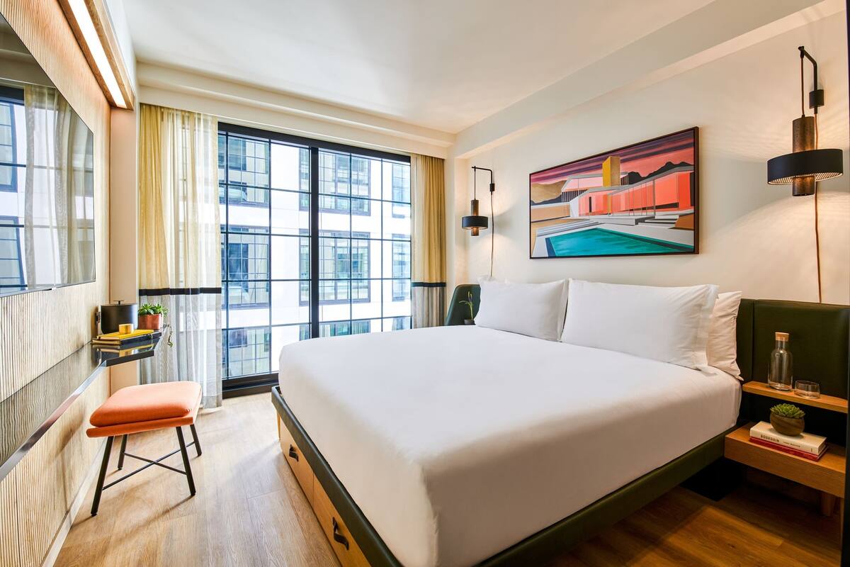 A hotel room with a large bed, a small stool, a nightstand, a painting, and a window with curtains.
